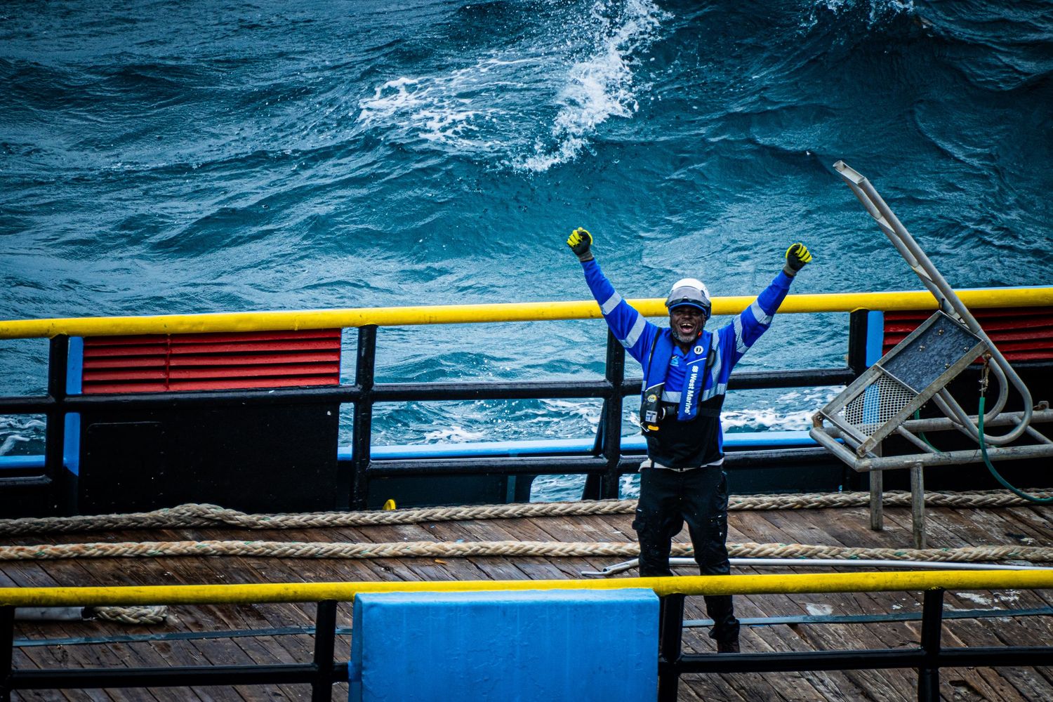 Worker in blue jacket raises fists triumphantly on offshore oil rig deck above rough ocean waves.