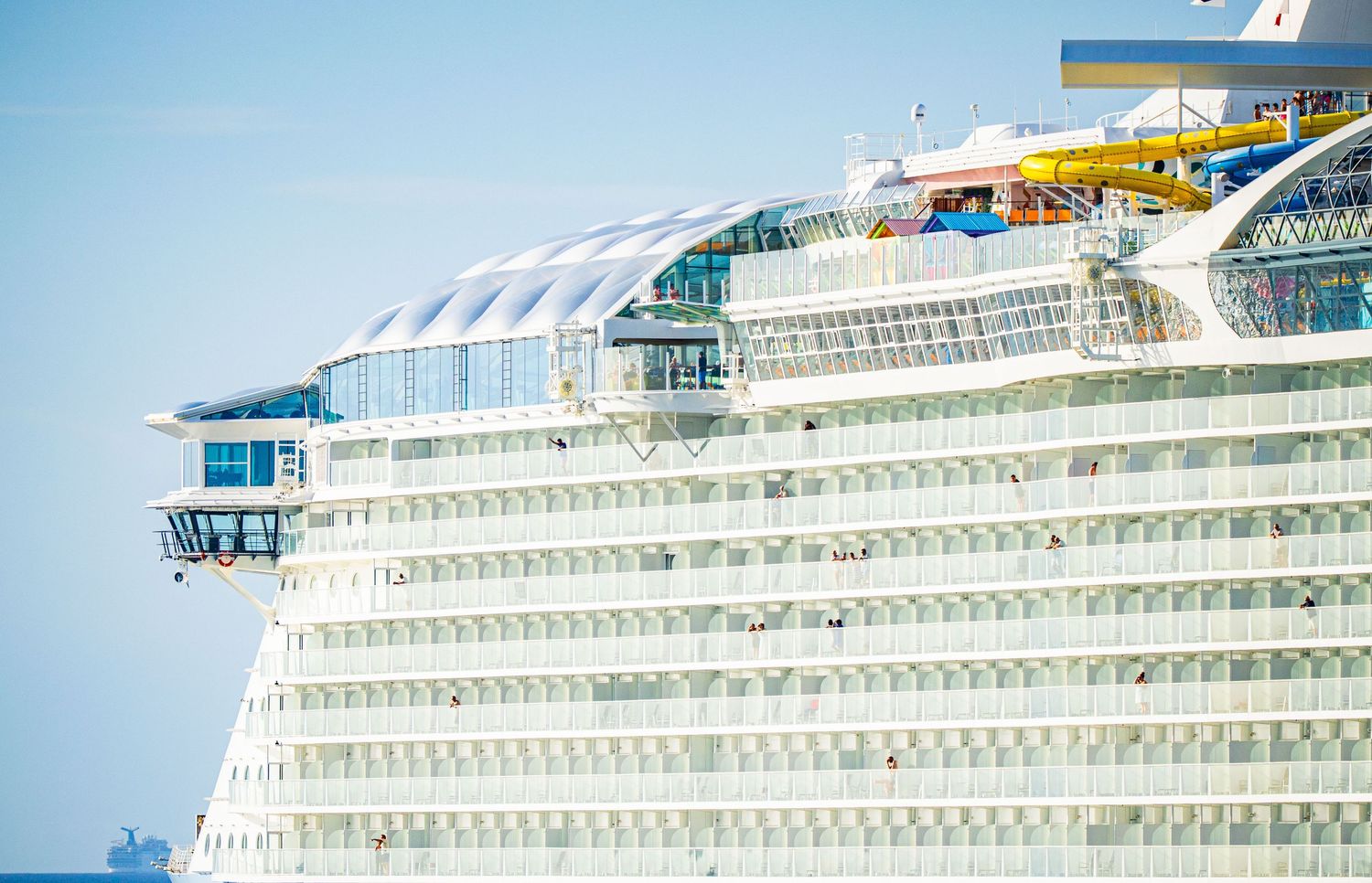 Large luxury cruise ship exterior showing multiple white decks, colorful water slides, and glass atrium under a clear blue sky.