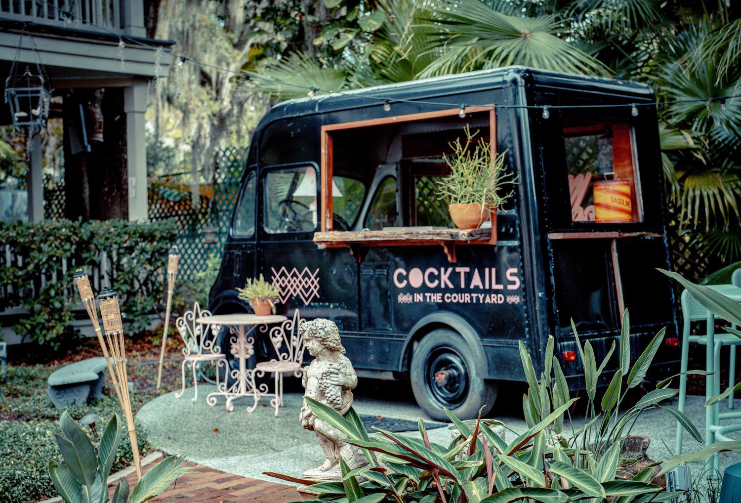 A vintage black truck parked in a lush tropical garden setting with palm trees and decorative lighting.