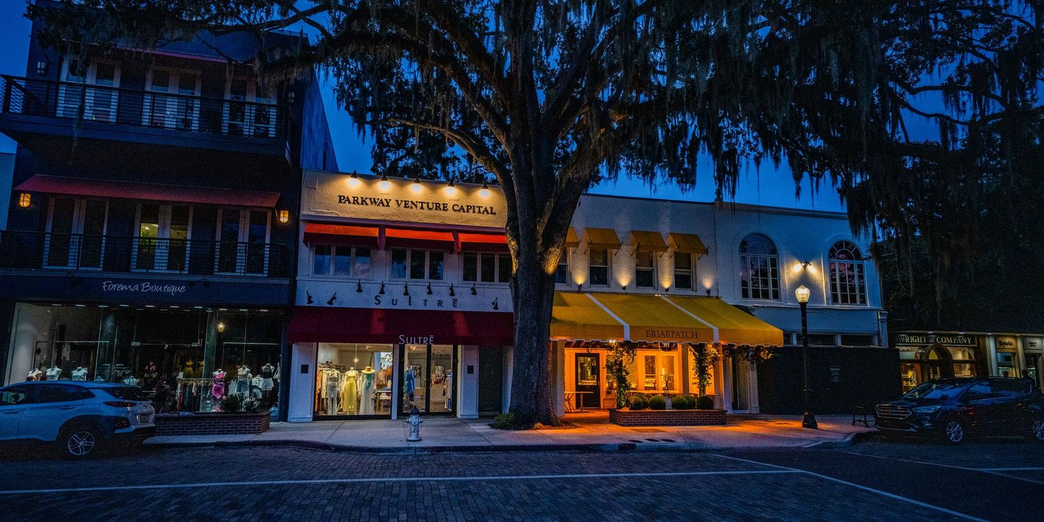 Row of illuminated storefronts and businesses along a downtown street at twilight.