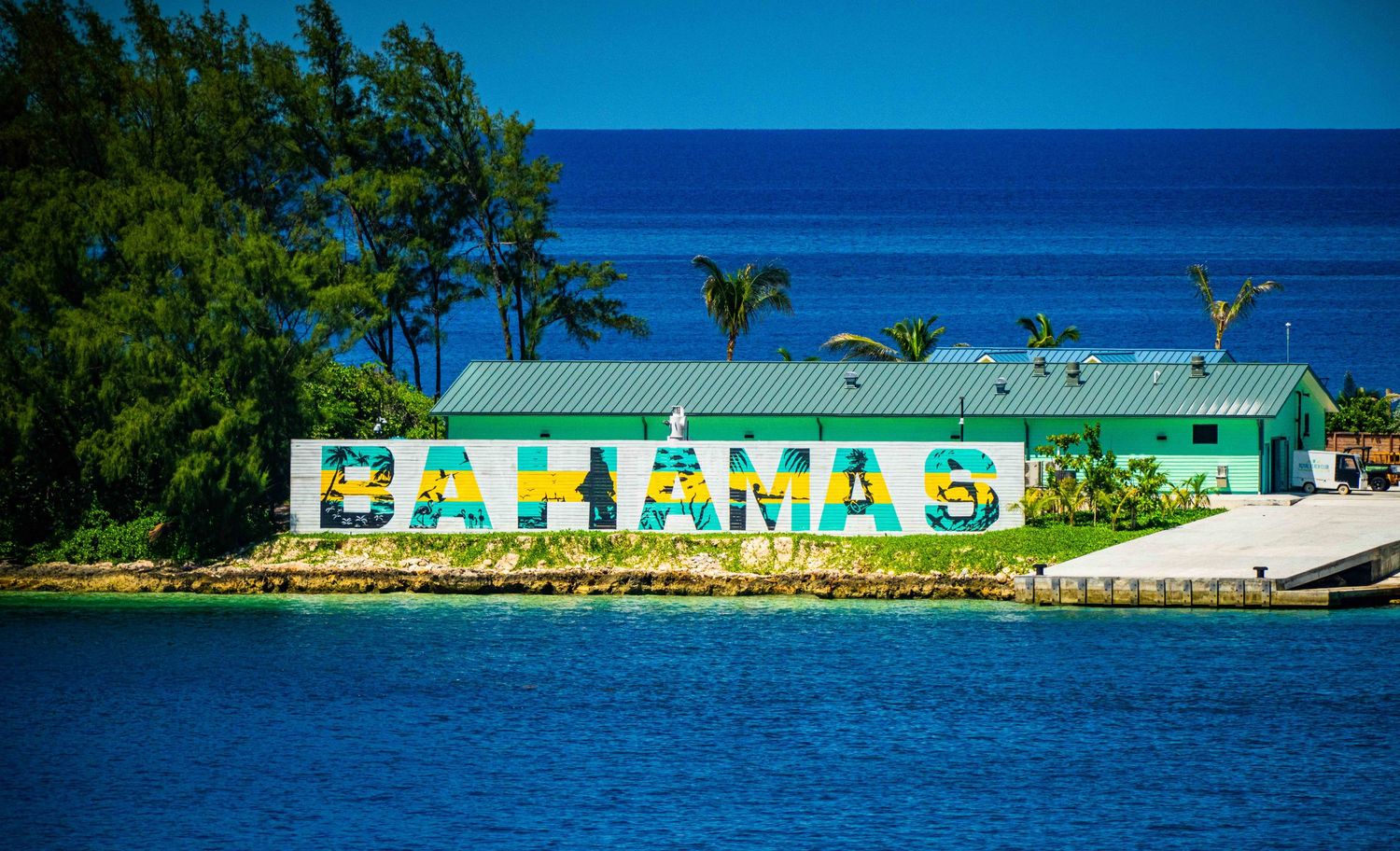 Colorful Bahamas sign painted on a waterfront building with lush green trees and vivid blue ocean in the background.