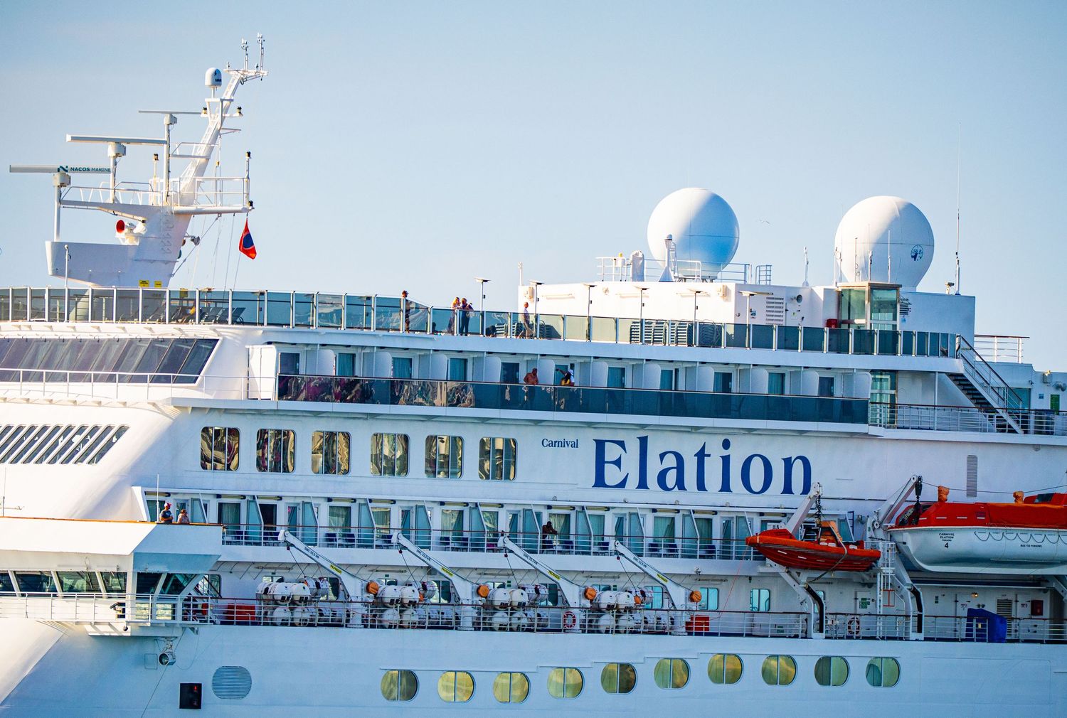 Carnival Elation cruise ship docked at port with passengers visible on multiple decks and a red lifeboat on the side.
