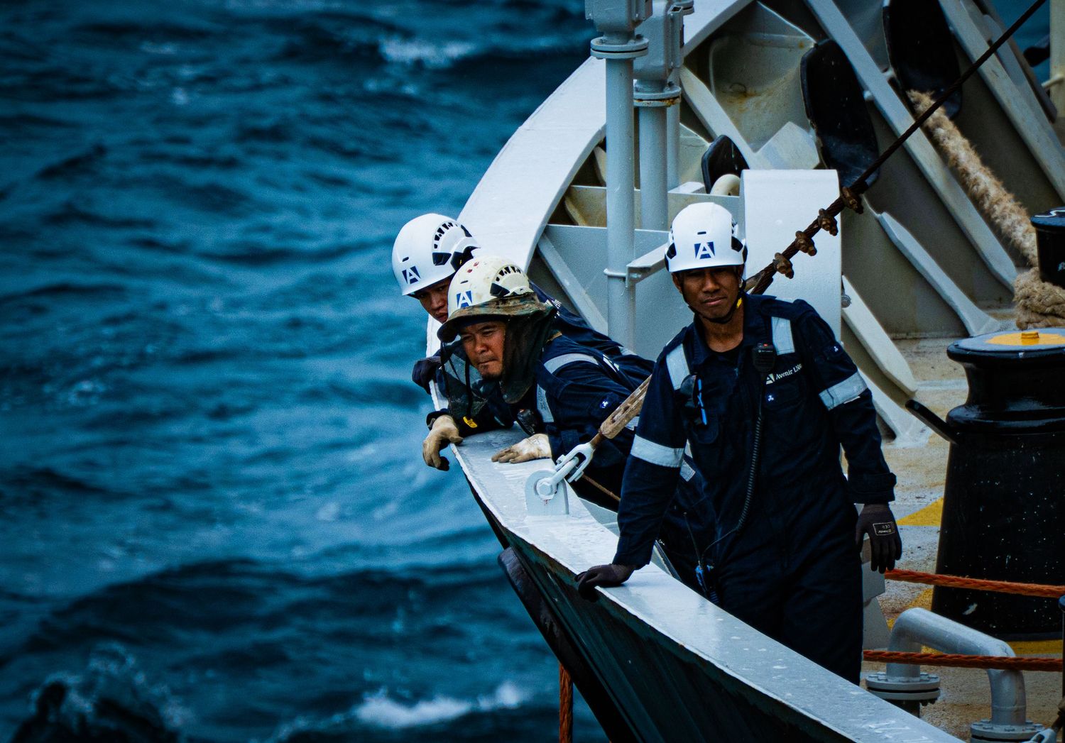 Two crew members in white helmets and navy coveralls working on a ship deck with rough ocean waves in the background.