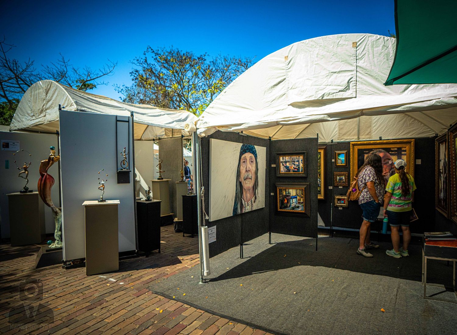 White tent gallery setup with artwork displayed on black walls under bright blue sky.