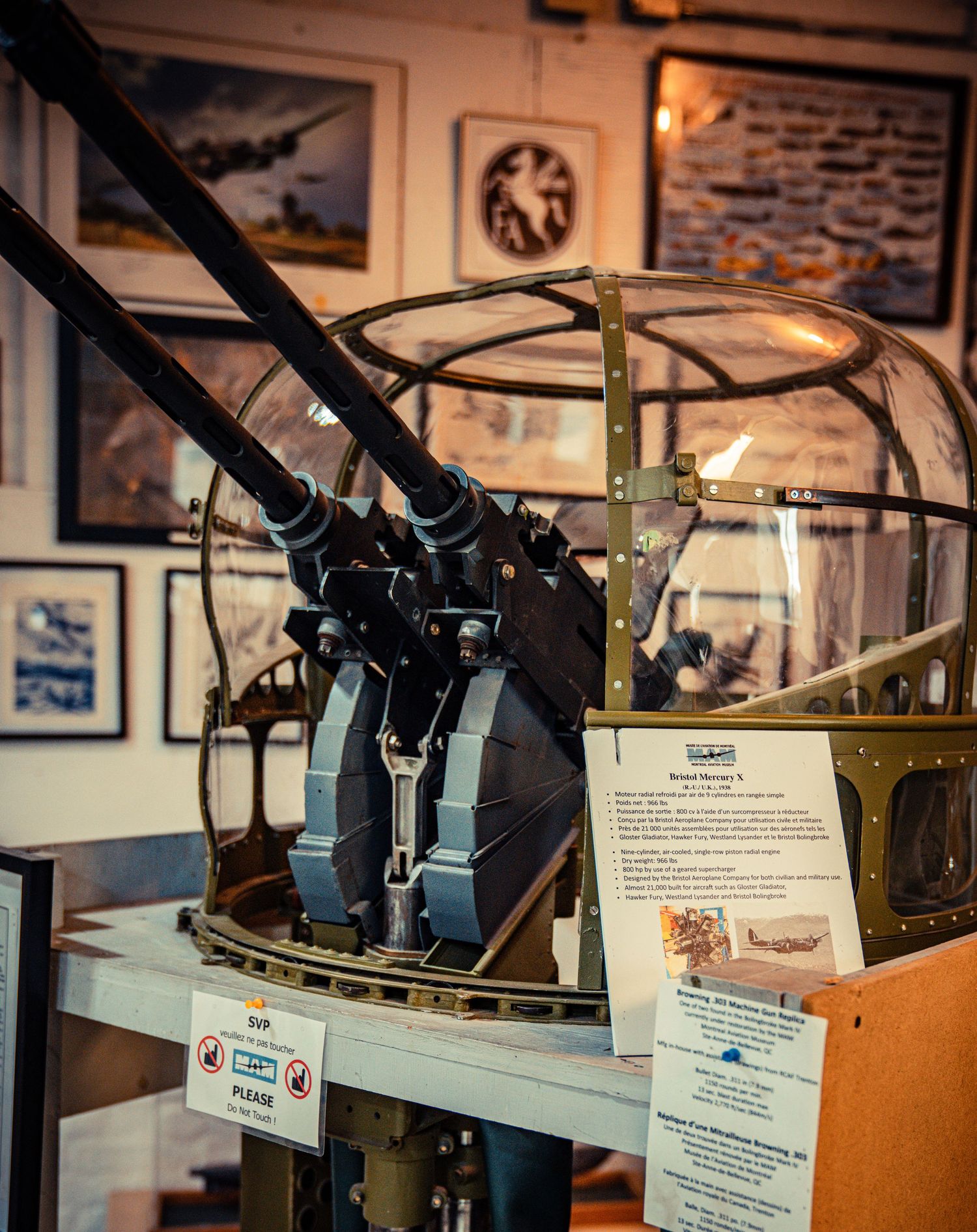 Historical aircraft gun mount display in a museum with framed aviation photos on the wall.