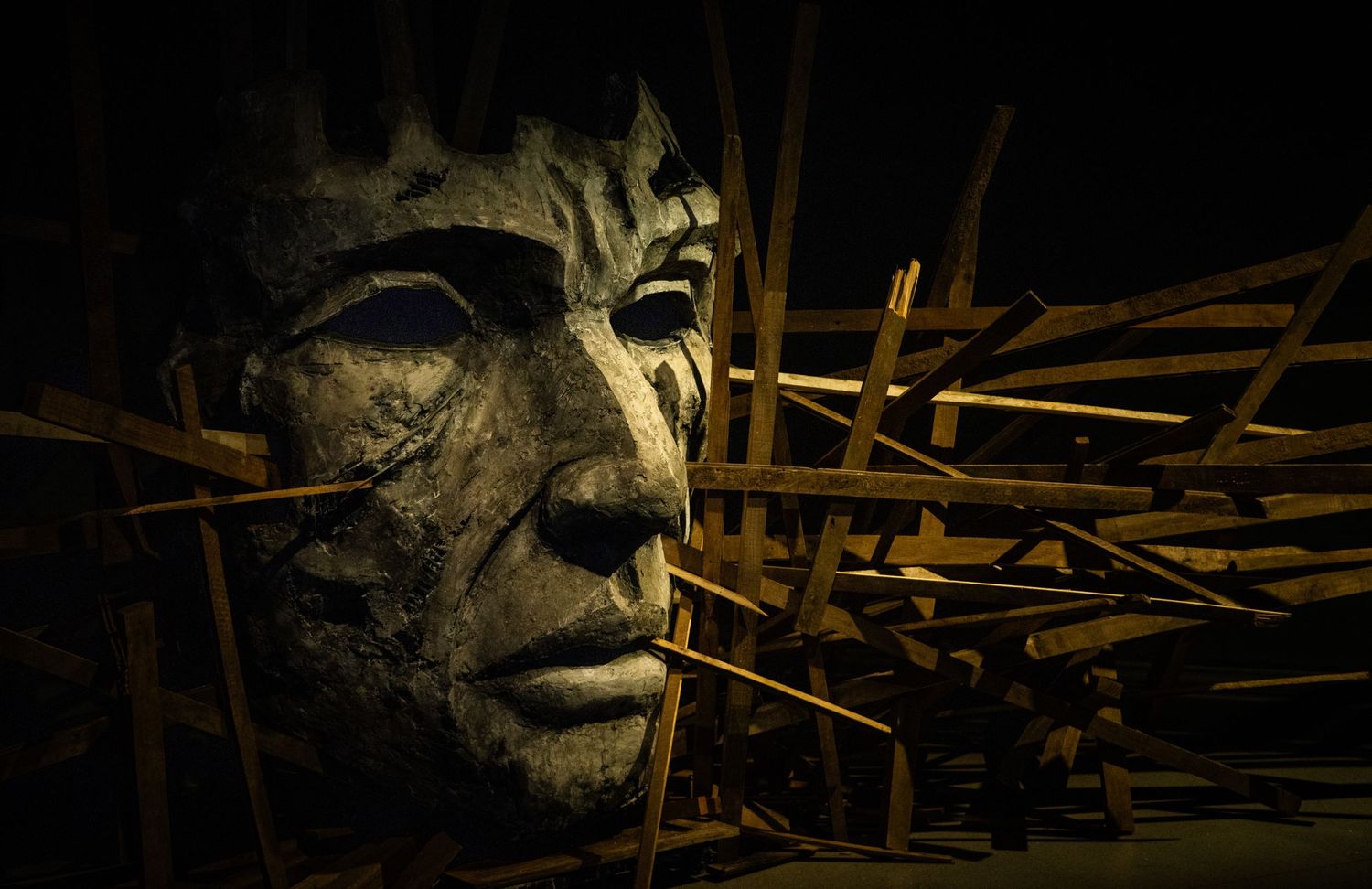 Dramatic sculpture with wooden sticks piercing through a stone face in dark lighting.