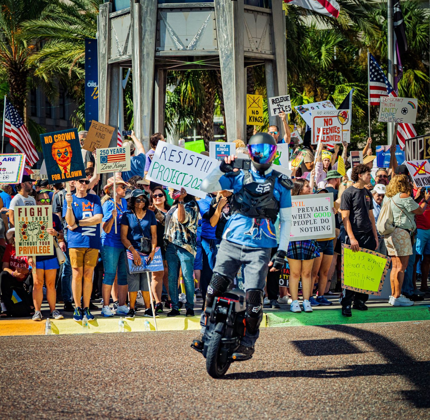 Large protest crowd gathers along palm trees with American flags and protest signs.