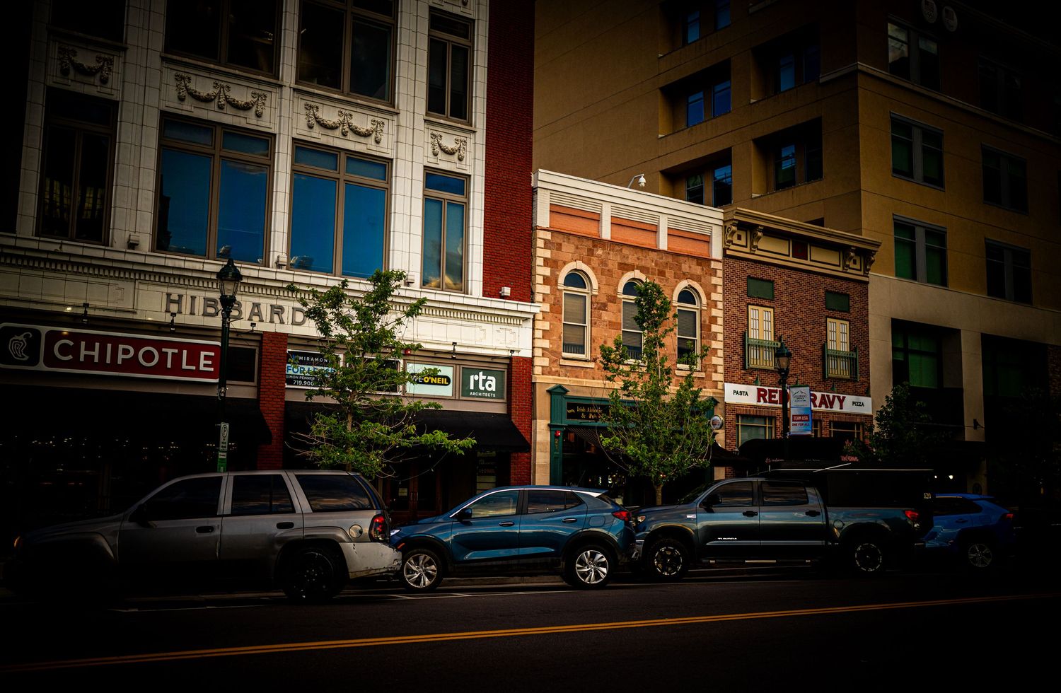 Downtown street with Chipotle and historic brick buildings, parked cars, and trees lining the sidewalk.