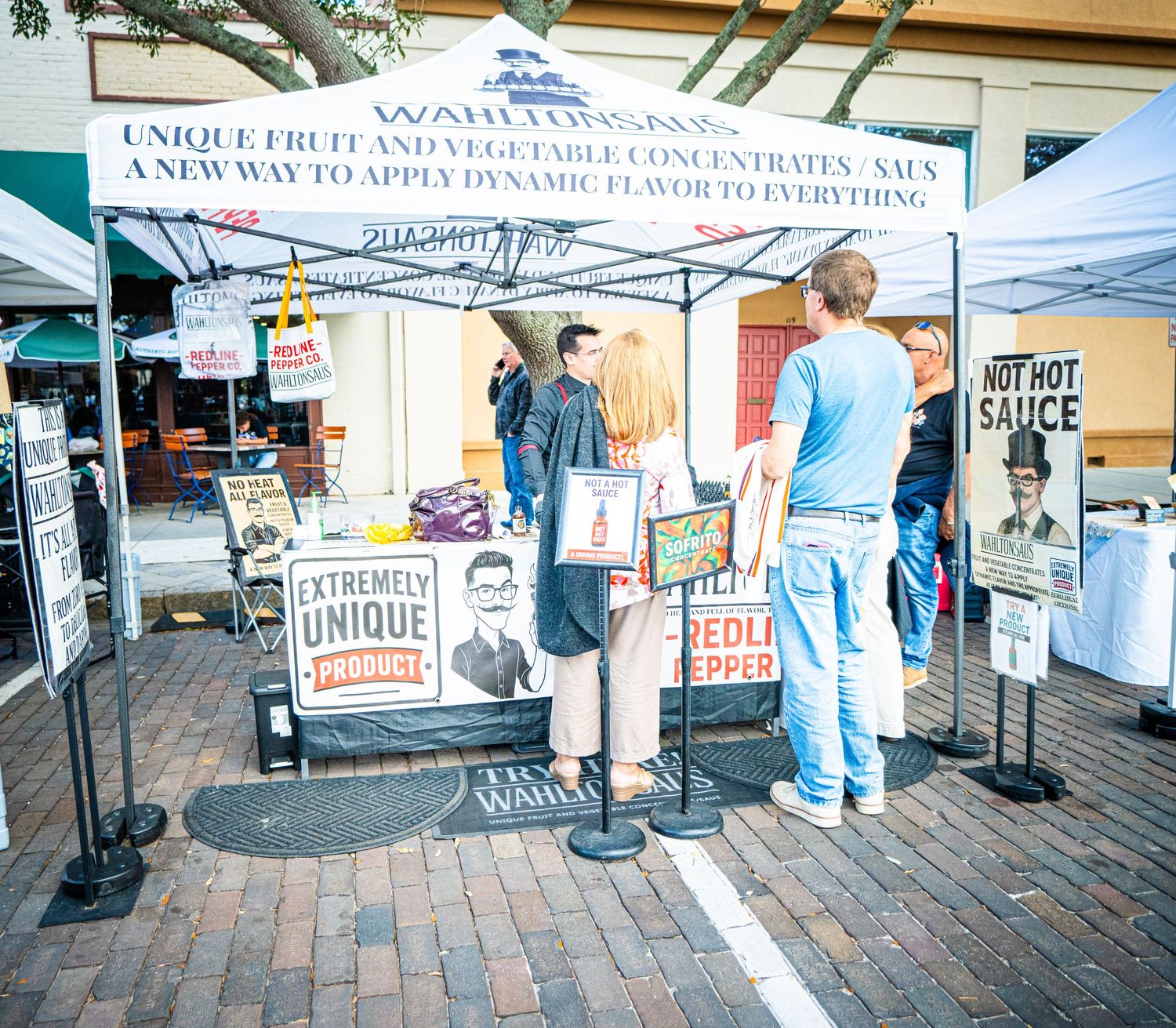 Panoramic view of vendors and white tents at an outdoor farmers market on a brick paved street.