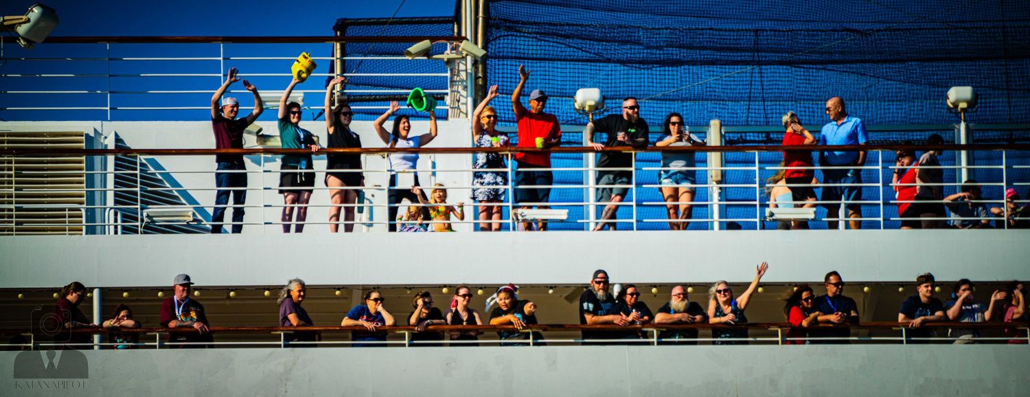 Cruise ship passengers gather along multiple decks of blue railings to observe something below.