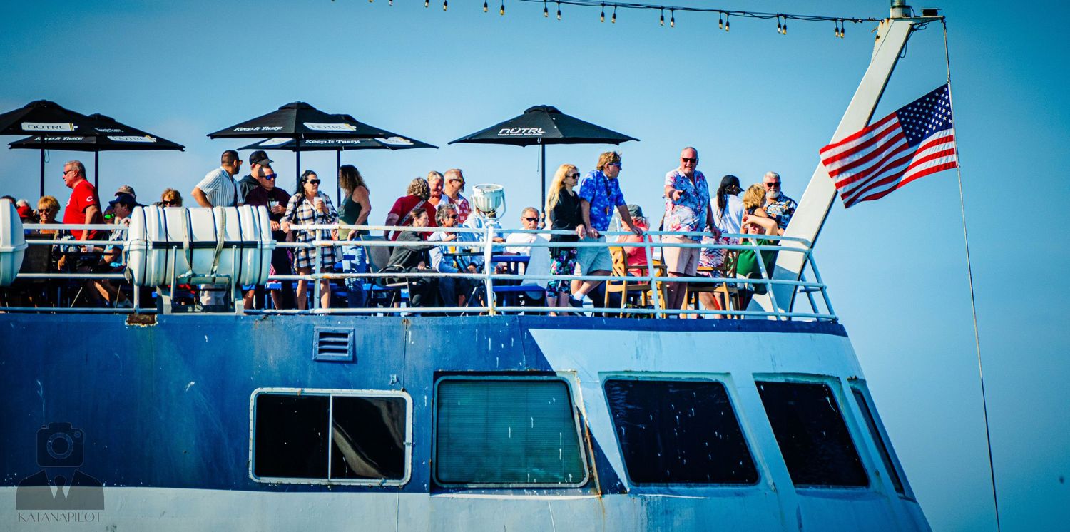 Panoramic view of a crowded boat deck with umbrellas and American flag against bright blue sky.