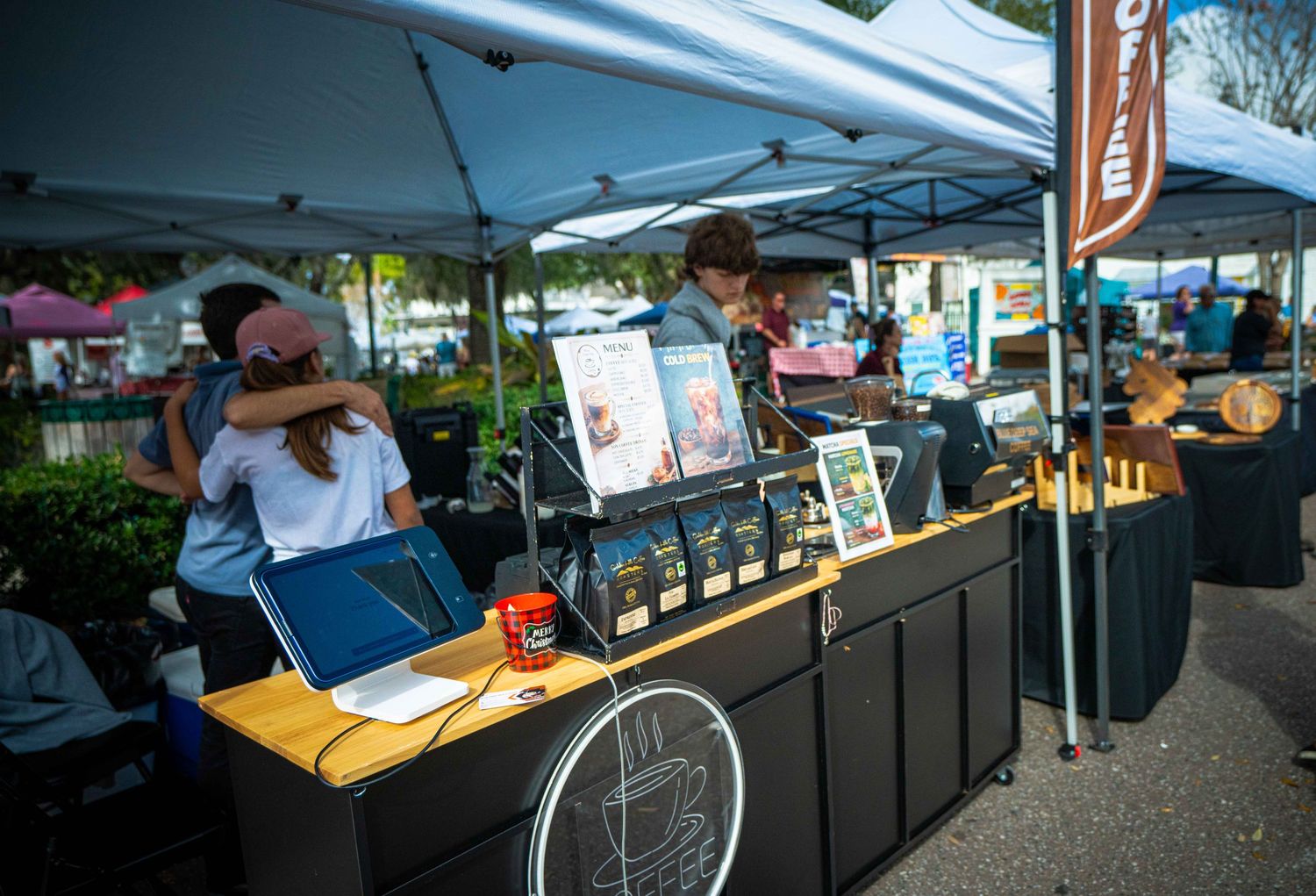Outdoor farmers market vendors display their products under white canopy tents on a sunny day.