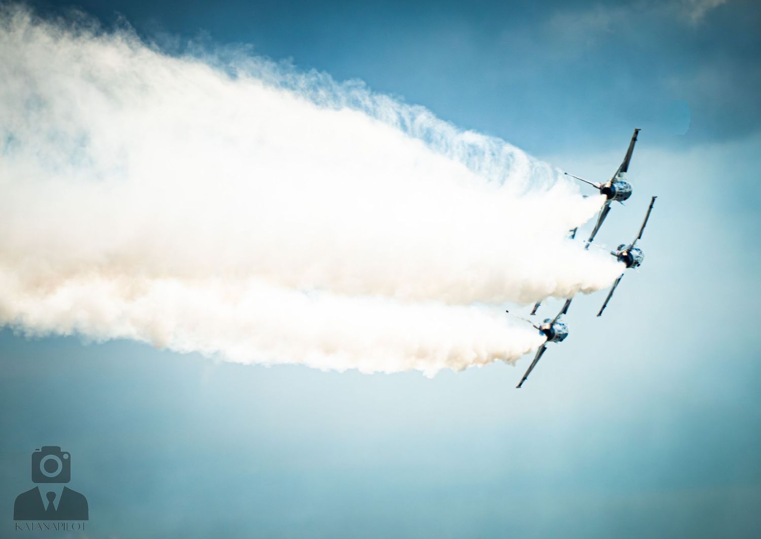 Four aircraft fly in tight formation at an airshow, trailing white smoke against a blue sky.