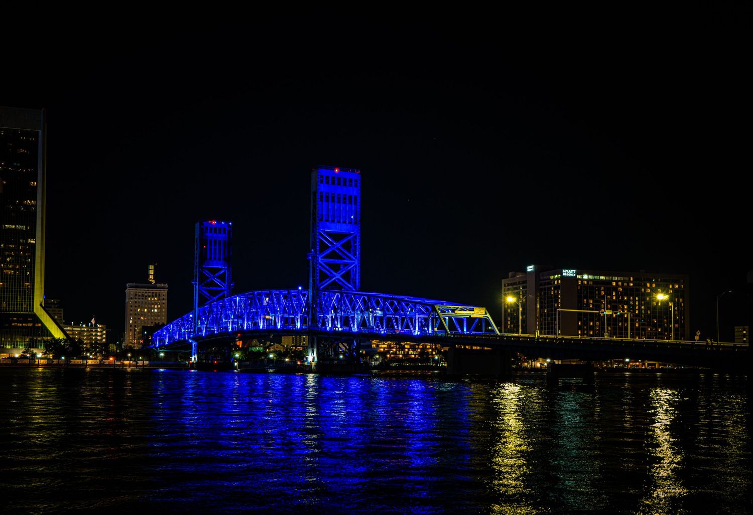 Blue-lit drawbridge reflecting on calm river at night with city skyline in background.