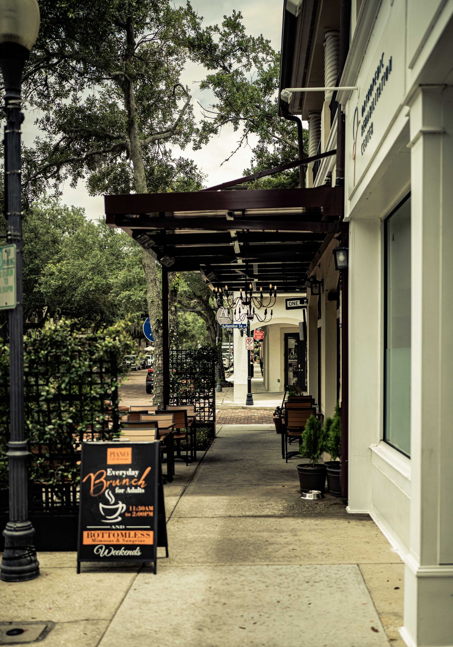 Charming outdoor sidewalk cafe with a chalkboard brunch sign, string lights, and lush trees lining a quaint downtown street.