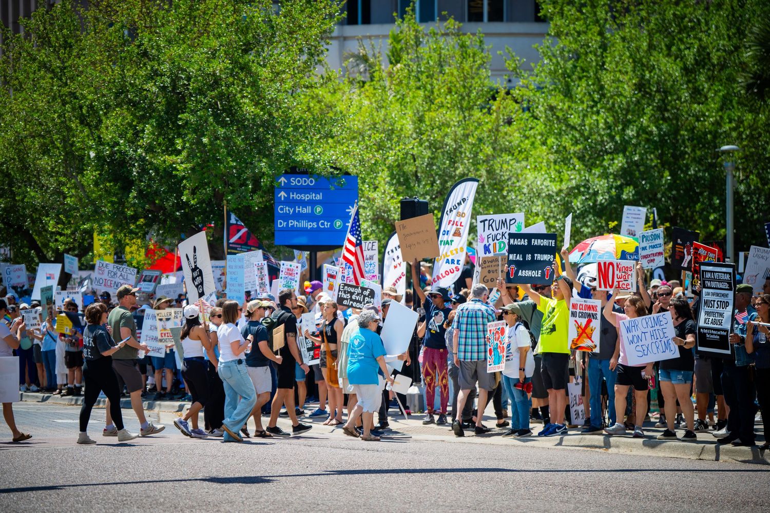 Large group of protesters holding signs gather on a tree-lined street during a peaceful demonstration.