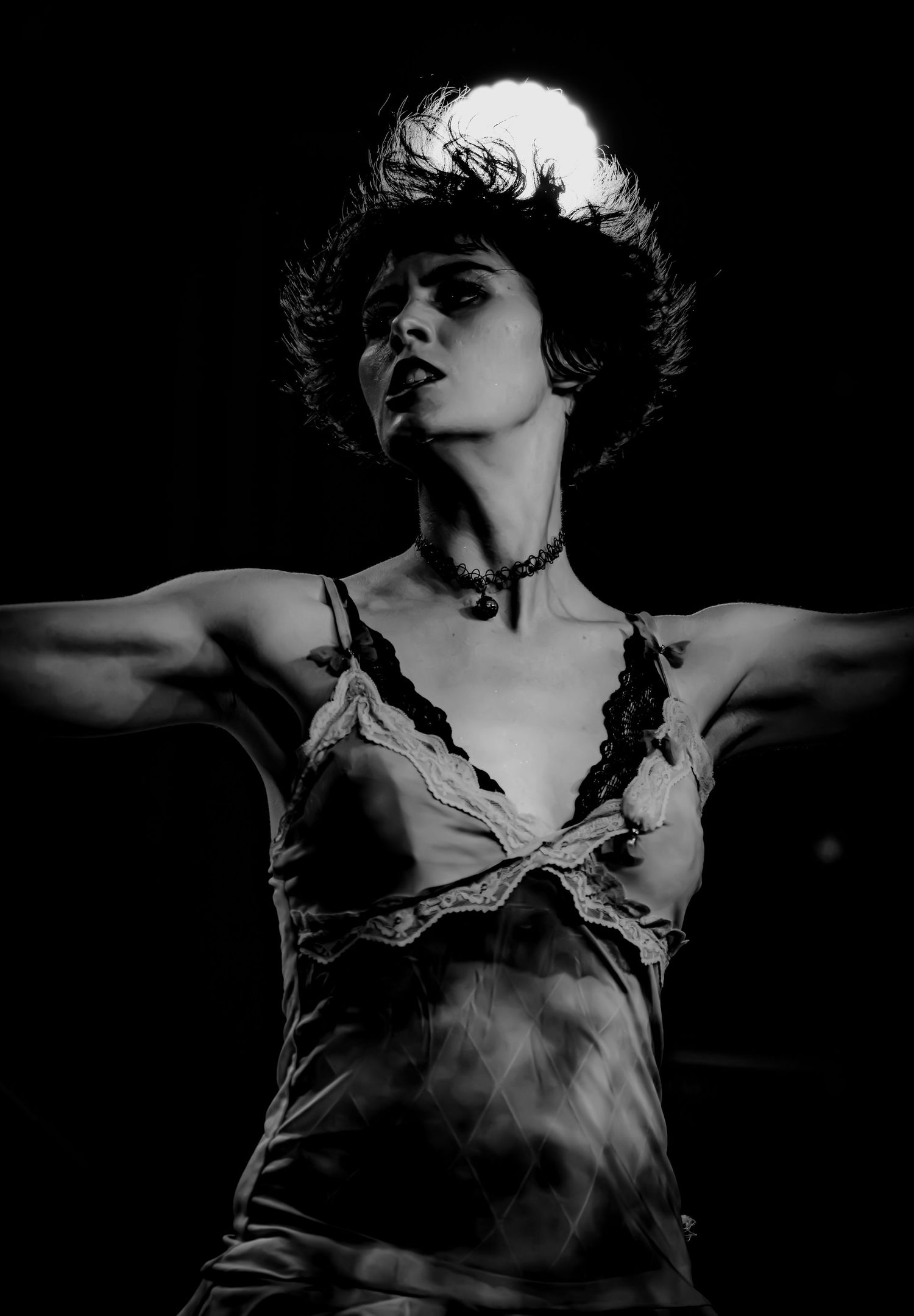 Black and white dramatic portrait of a woman in lace slip dress with choker, arms outstretched under spotlight.