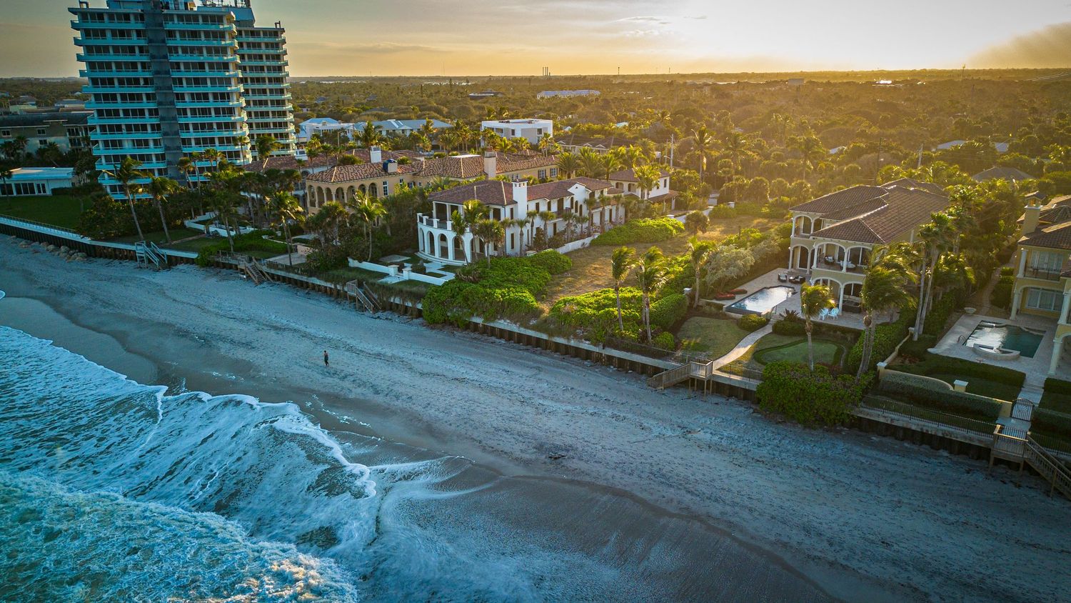 Drone shot of luxury beachfront homes and a high-rise building along the shoreline at dusk.