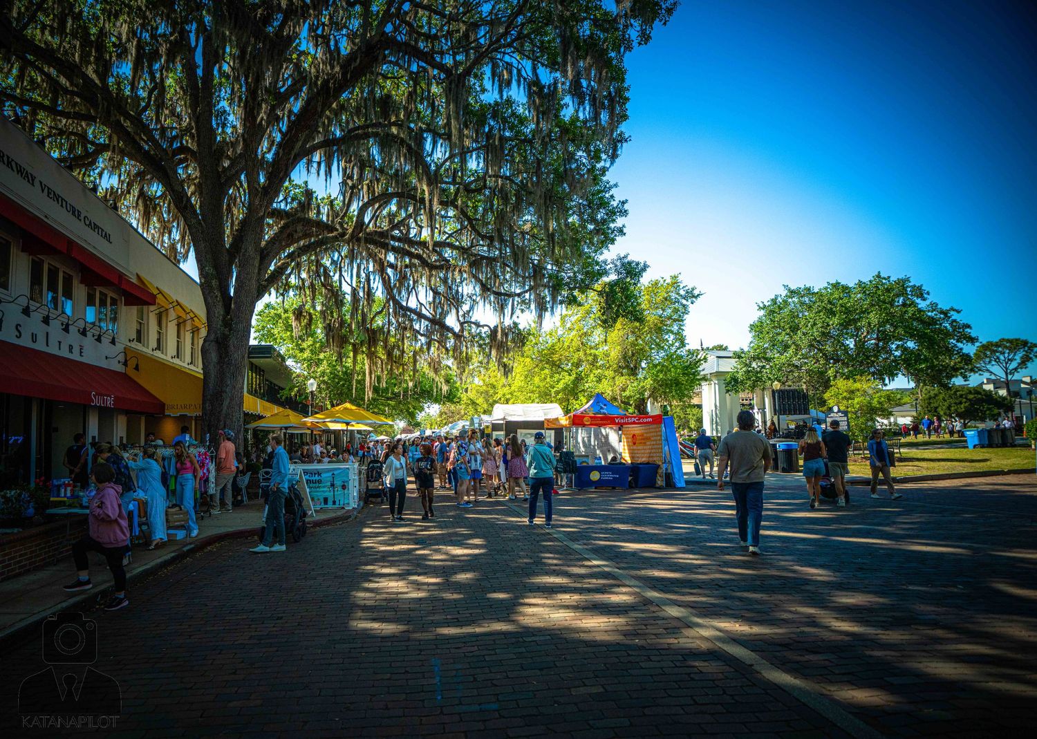 Wide panoramic view of a shaded outdoor marketplace with trees and blue sky overhead.