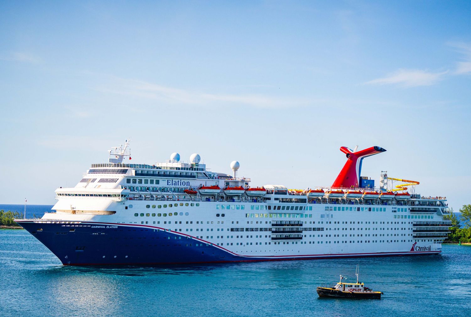 Large Carnival cruise ship sailing through blue waters near a tropical port destination.