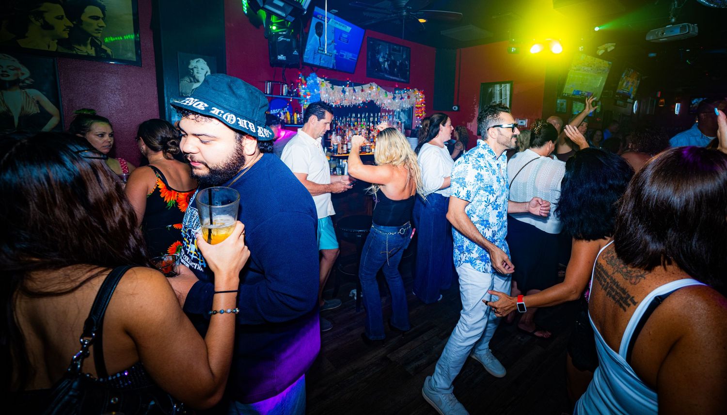Lively nightclub scene with people dancing and socializing under purple and yellow lights.