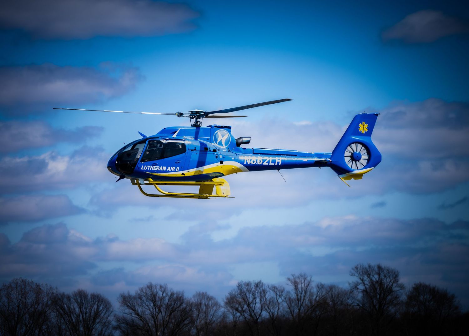 Blue and yellow helicopter flying against cloudy blue sky.