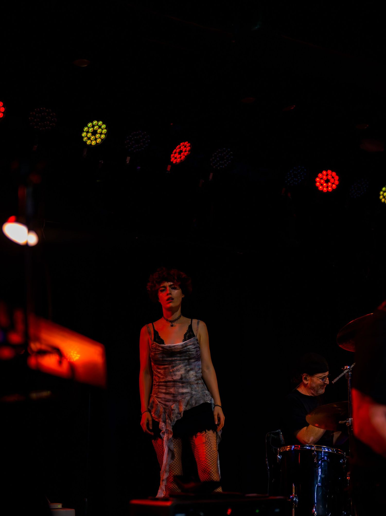 Female performer on dark stage with colorful overhead lights, wearing mini dress, with drummer visible in background.
