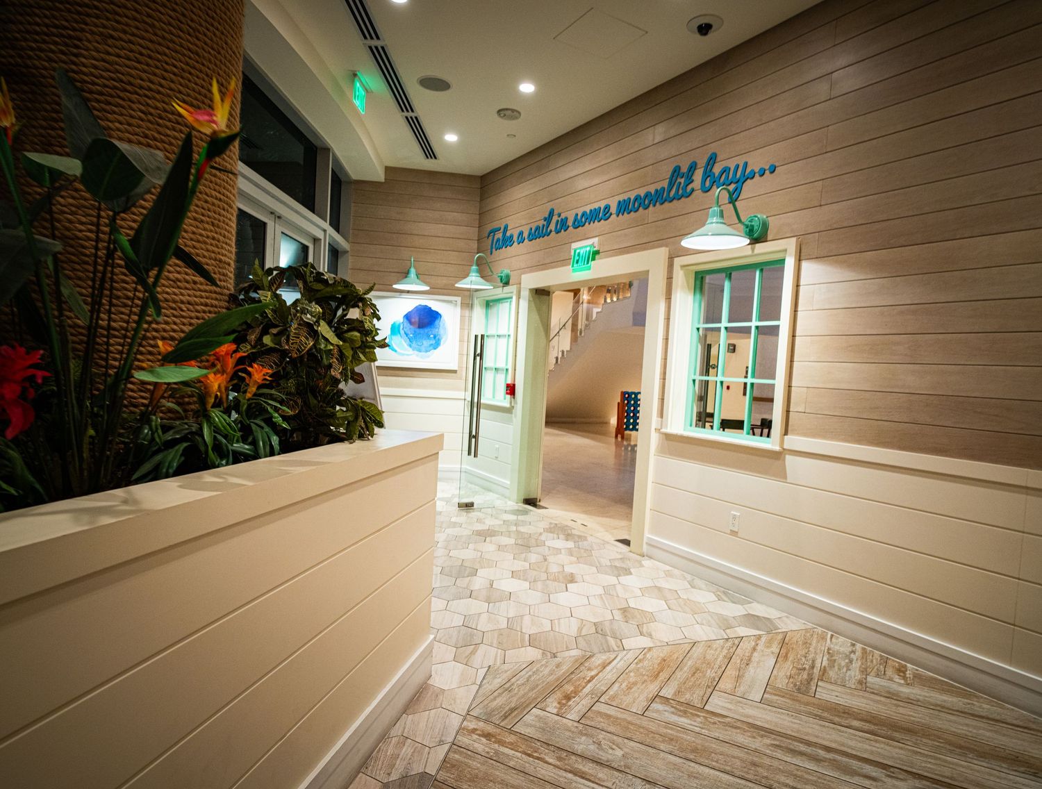 Bright coastal-themed hotel corridor with wood-paneled walls, herringbone floors, and teal pendant lights.