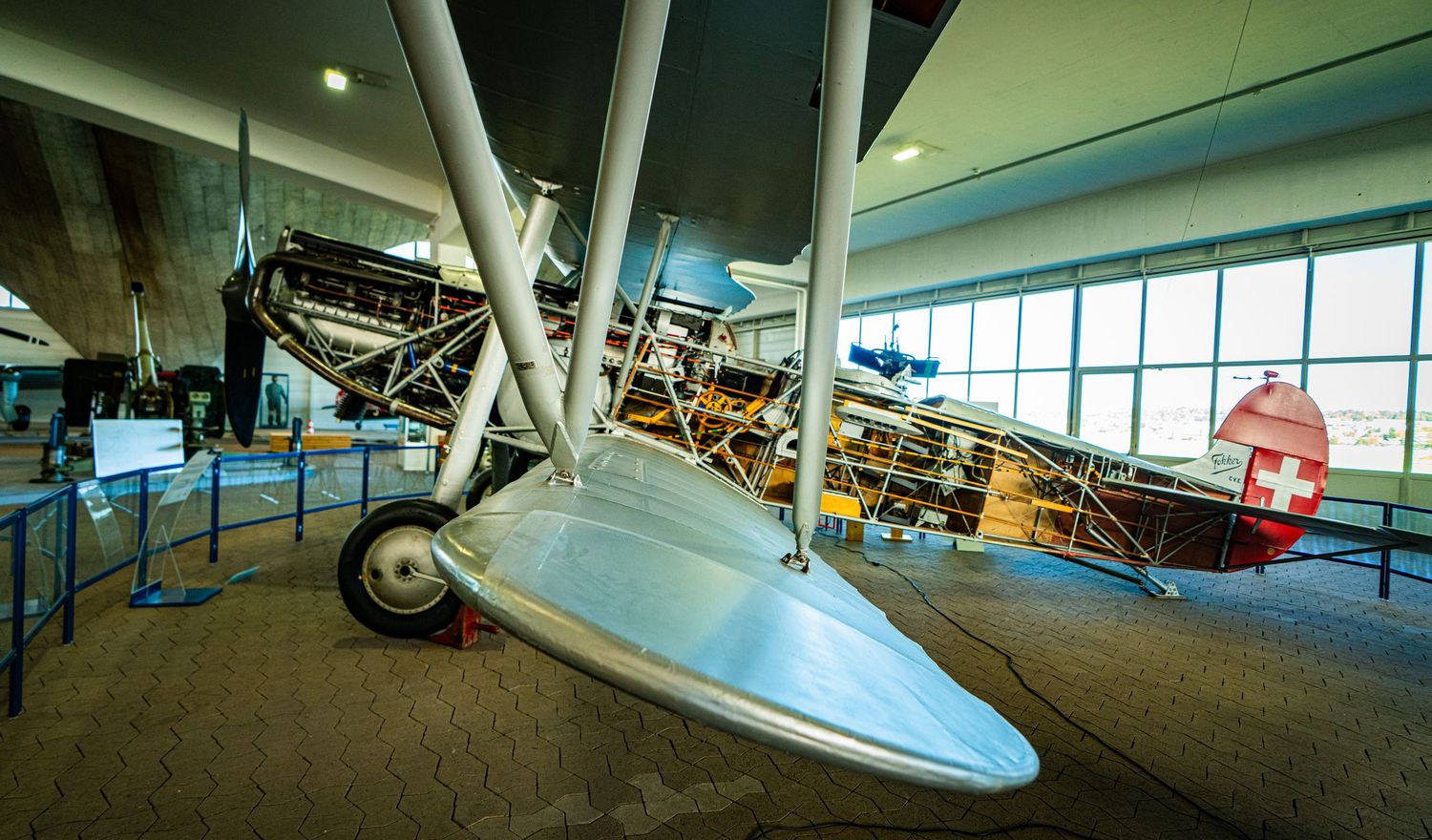 Vintage biplane with silver wing and exposed struts on display in a bright aviation museum, with a Swiss flag emblem on the tail.