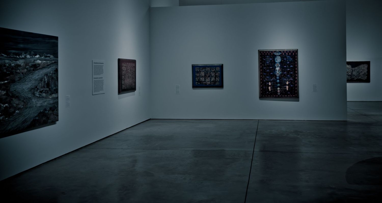 Dimly lit art gallery with dark paintings displayed on white walls and polished concrete floors.