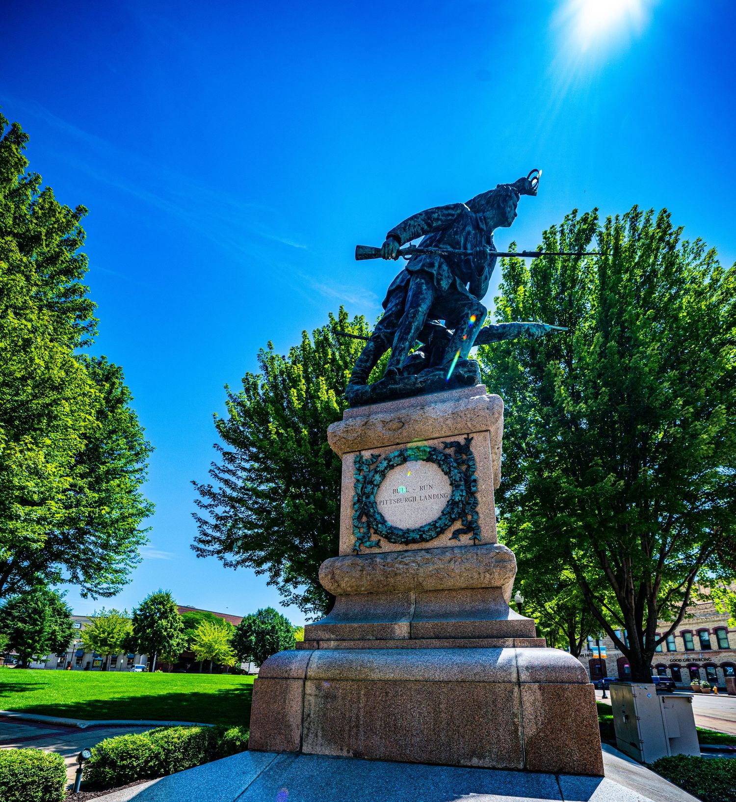 Bronze war memorial statue against bright blue sky with trees in a park setting.
