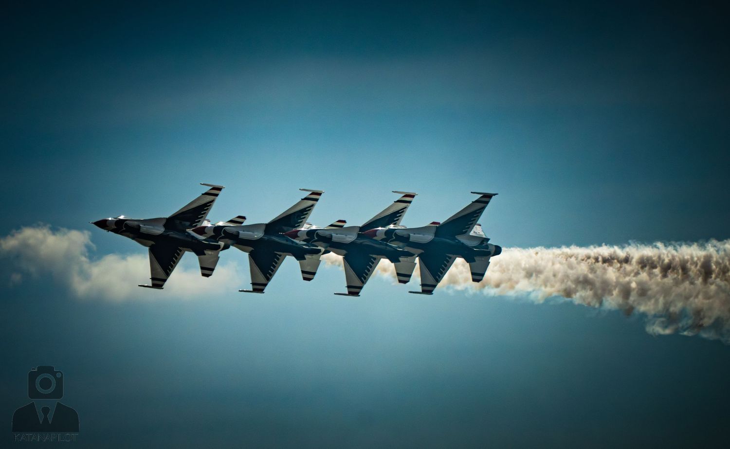 Four U.S. Air Force Thunderbirds fly in tight diamond formation, trailing white smoke against a vivid blue sky.