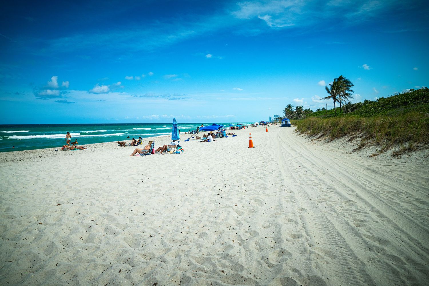 Beachgoers relax on a white sandy beach with blue umbrellas and ocean waves in the background.