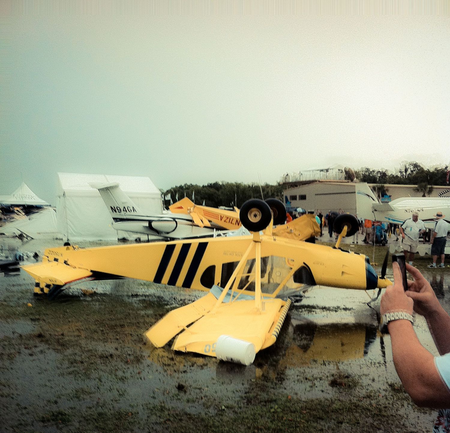 A vintage yellow training aircraft parked on a grassy field alongside other small planes at an airfield.