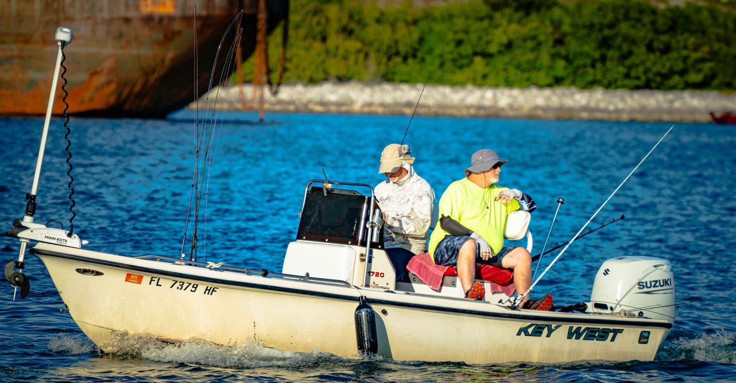 Two people fishing from a small white boat on calm blue waters near a lush green shoreline.