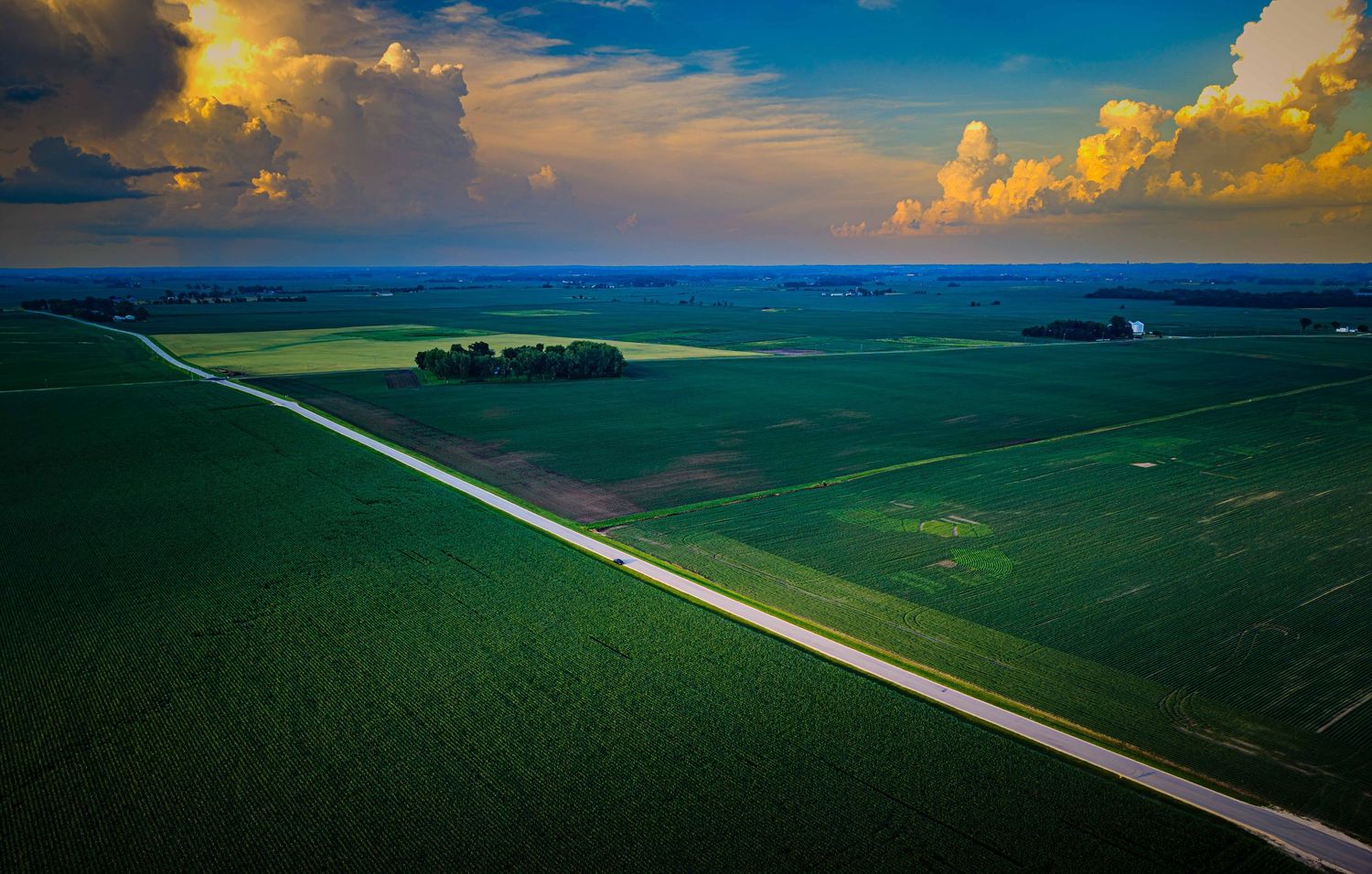 Aerial view of vast green farmfields with straight road under dramatic sunset clouds.