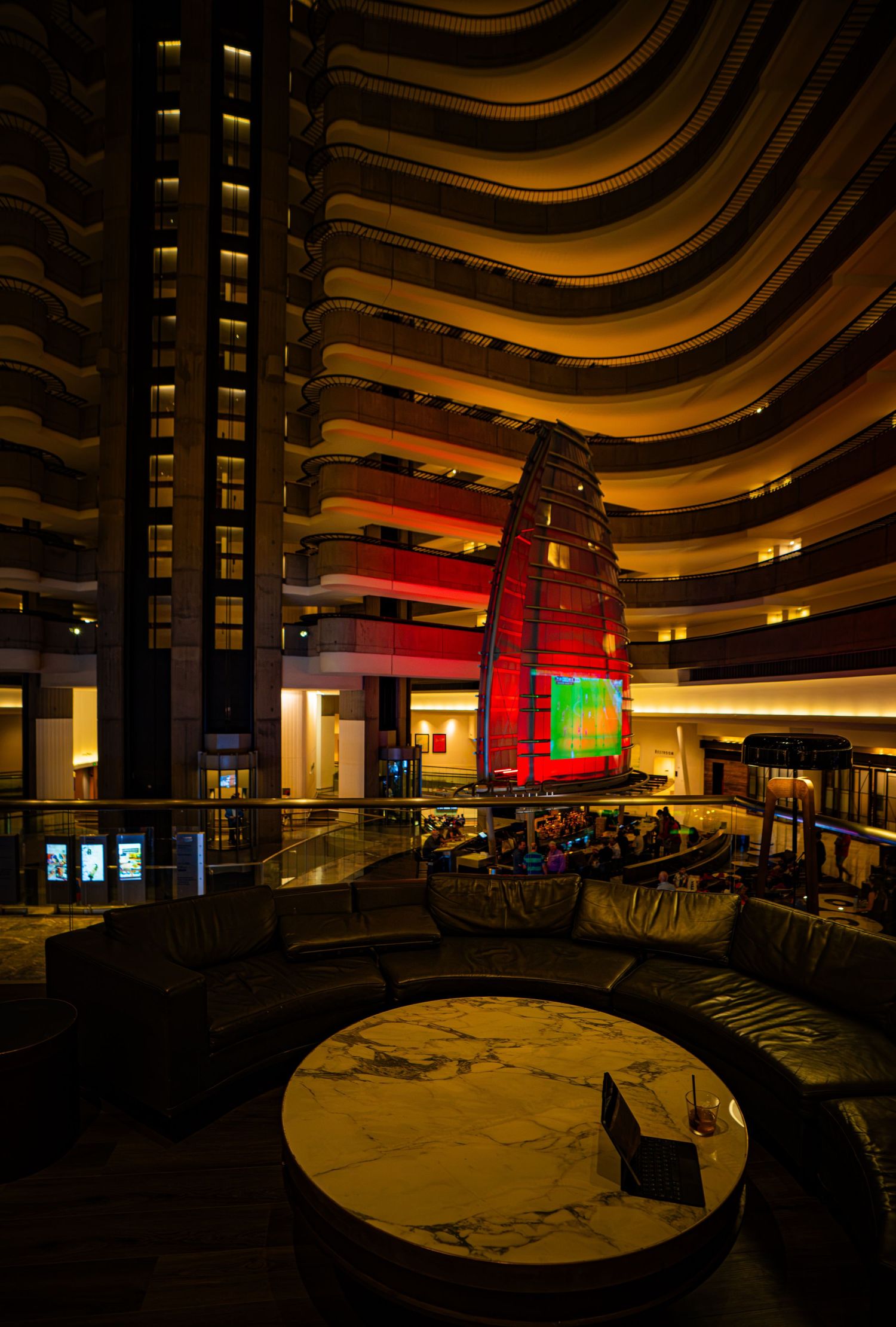 Grand hotel atrium lobby with curved balconies, glowing red sail sculpture, and marble table at night.