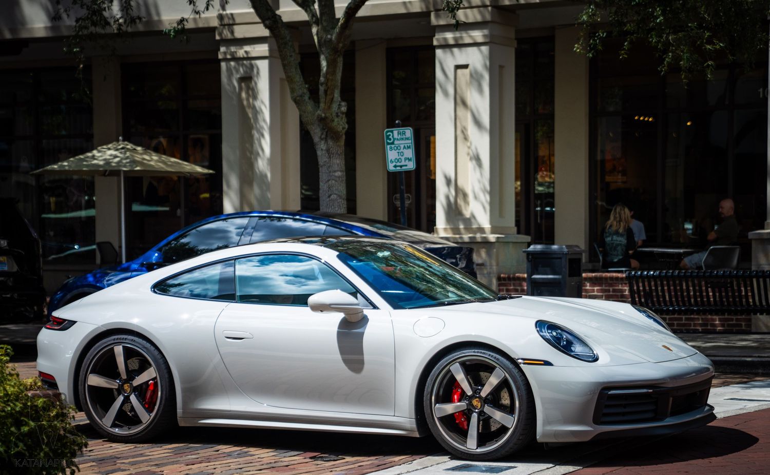 Luxury sports cars including Porsches in silver and blue are displayed along a brick paved street.