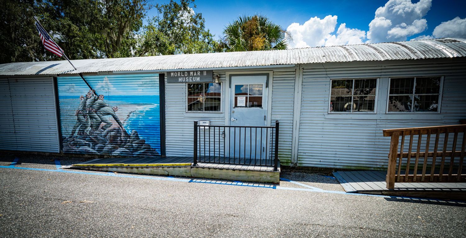 Long white building exterior with sidewalk and landscaping under blue sky.