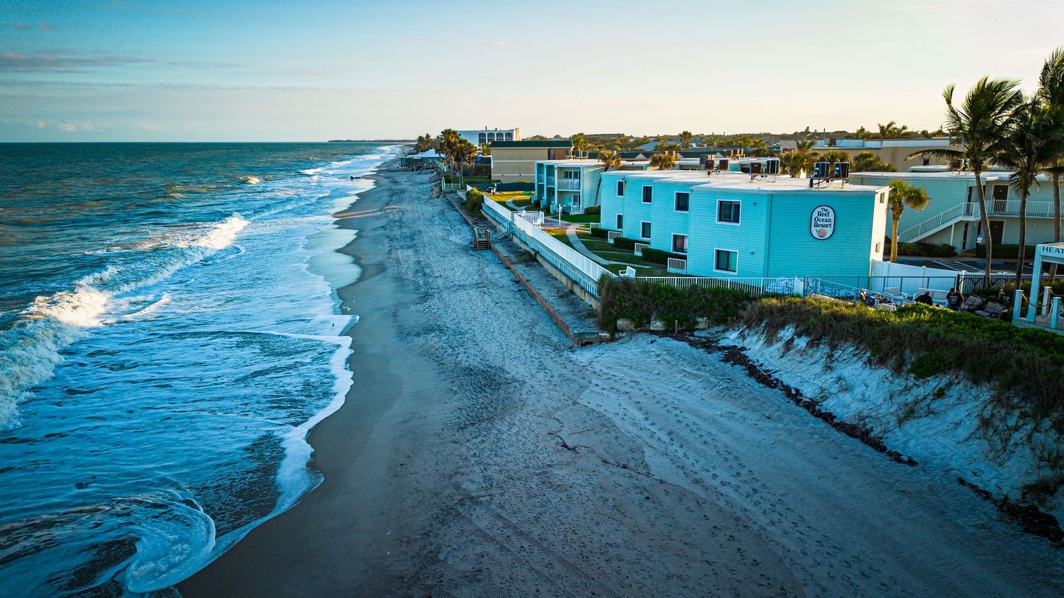 Coastal properties in vibrant blue colors line a beach with crashing waves at dusk.