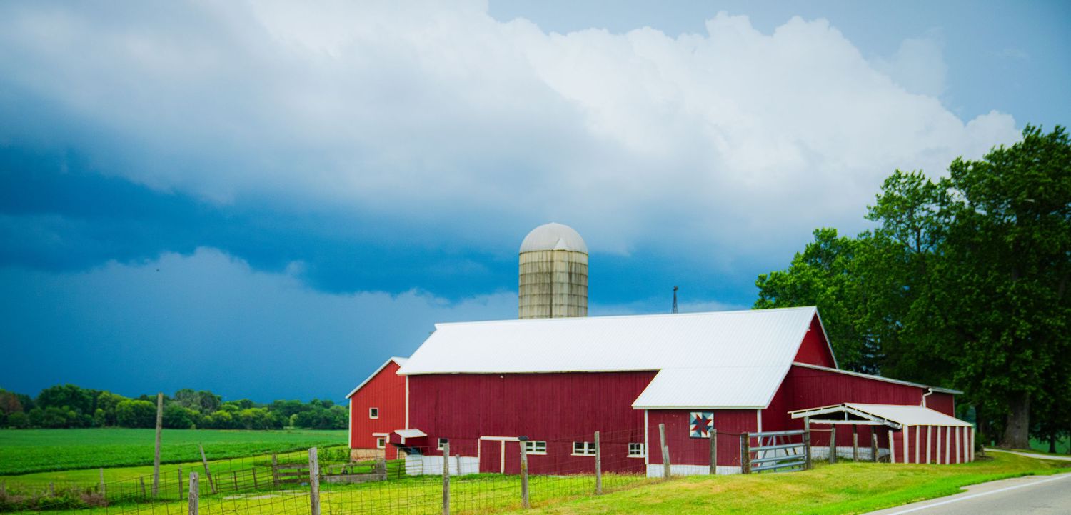 Rural landscape with red barns and farm buildings under dramatic cloudy skies.