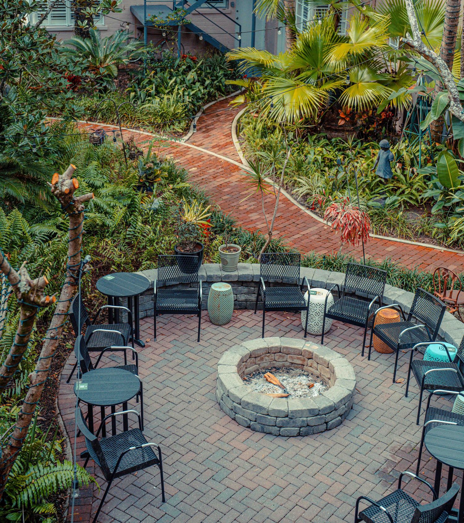 A circular brick paver patio features a stone fire pit surrounded by black metal chairs and lush tropical landscaping.