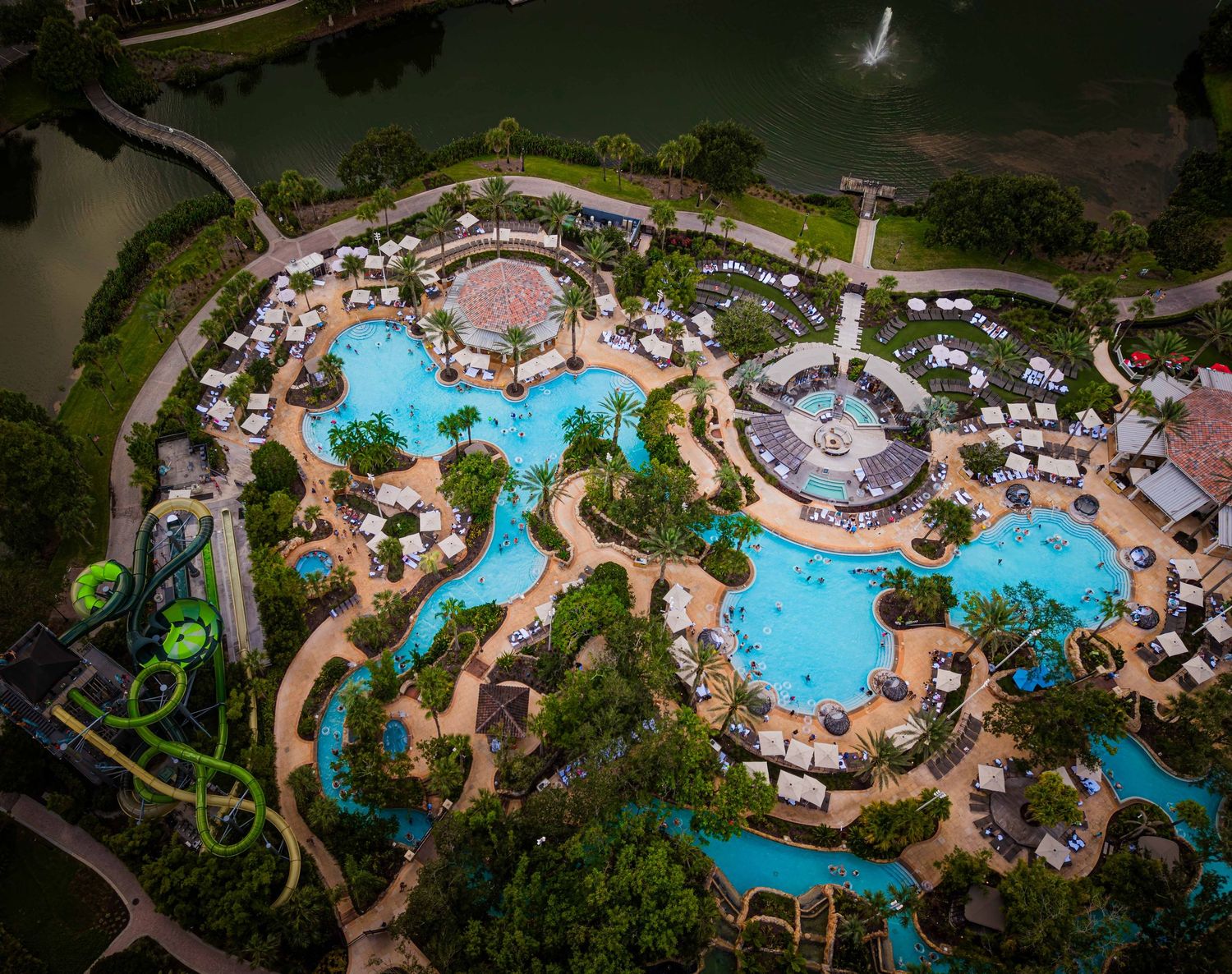 Aerial view of a luxury resort pool complex with winding water slides, lounge chairs, and a serene lake surrounded by lush trees.