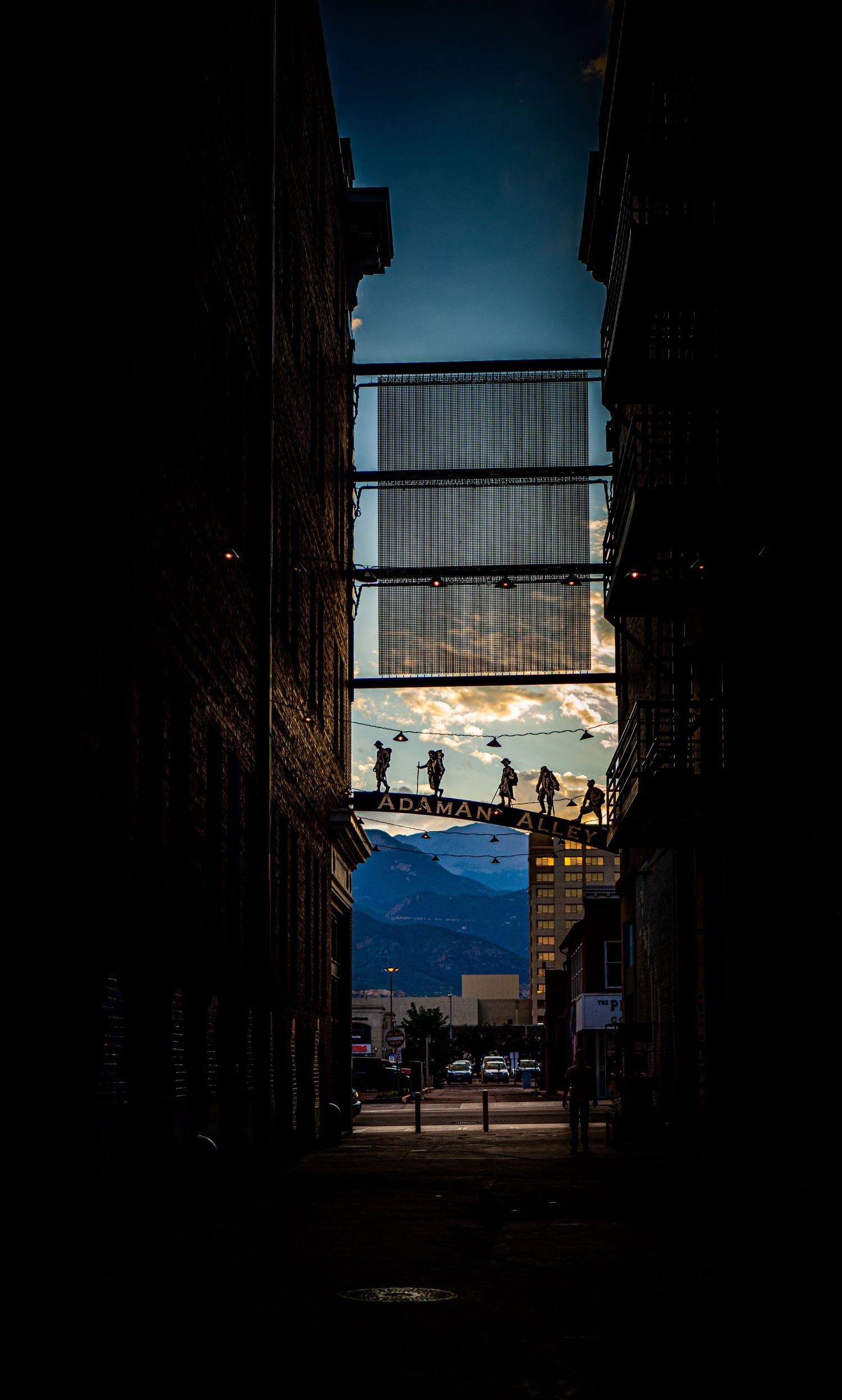 Dark alley leading to a dramatic sunset sky with silhouetted figures on a sign above a city street.