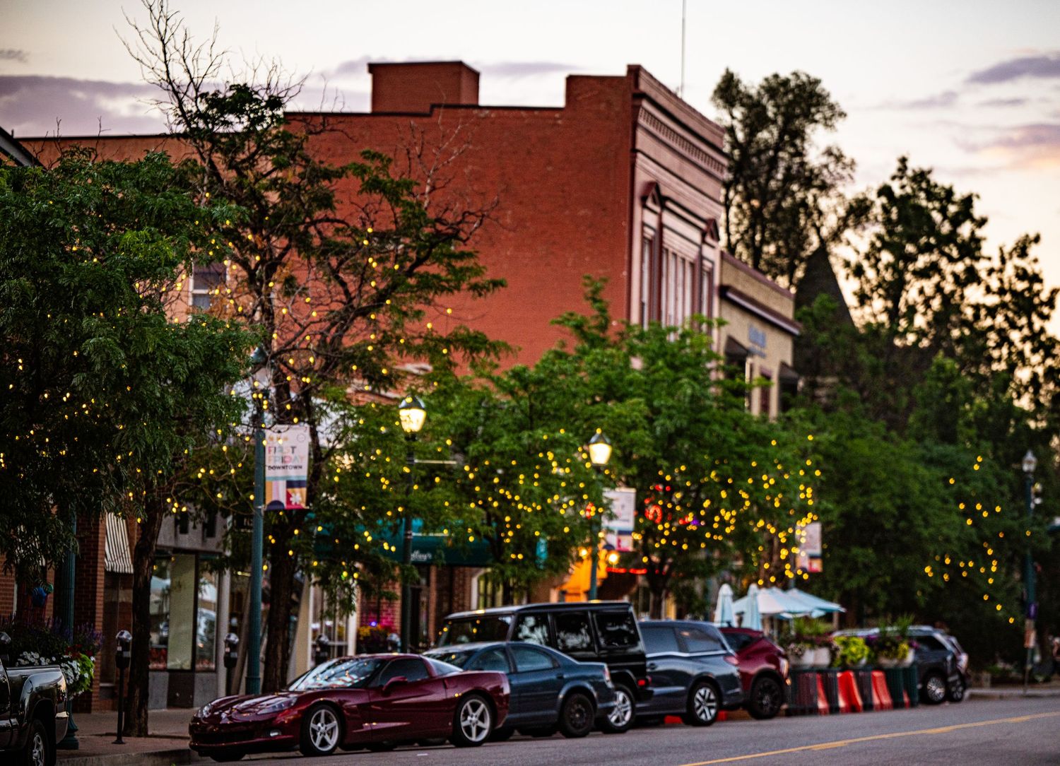 Charming small-town main street at dusk with string lights, parked cars, and brick buildings surrounded by lush green trees.