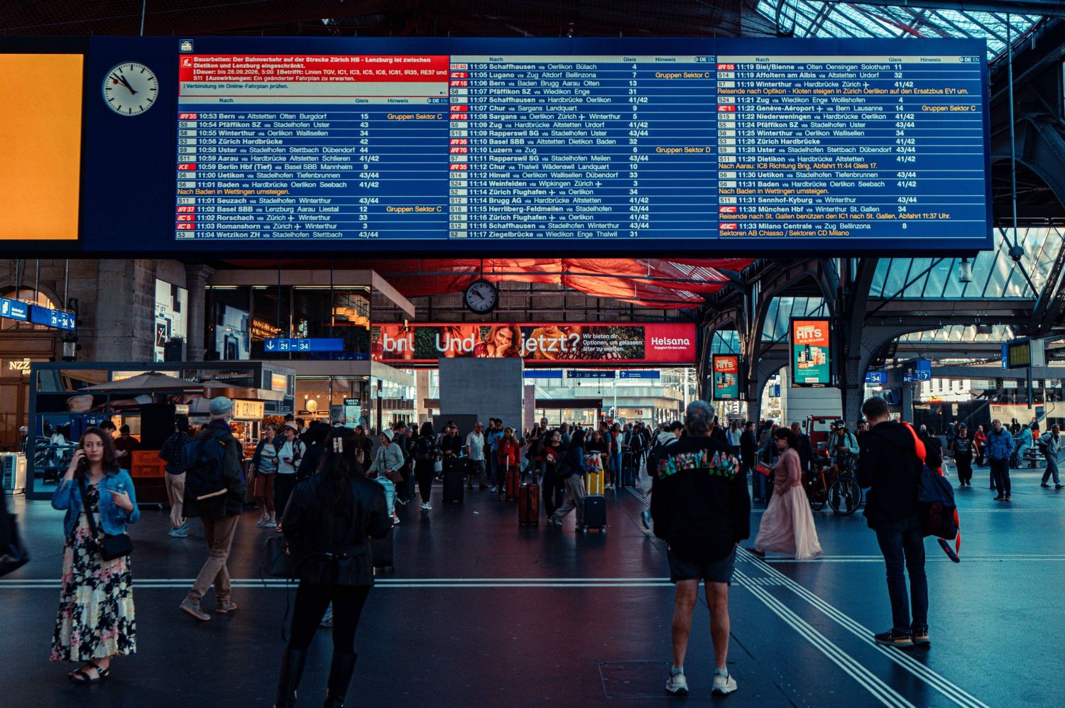 Crowded train station concourse with travelers walking beneath a large blue departure board and overhead clock at dusk.