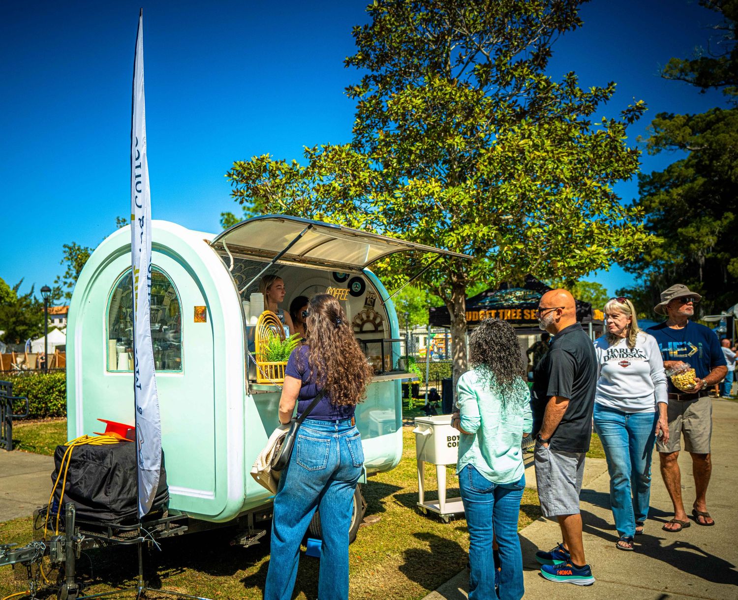 Row of vintage travel trailers and food vendors at an outdoor event with attendees browsing under blue skies.