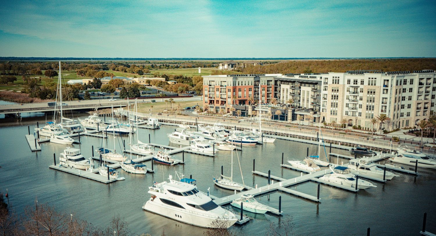 Aerial view of a modern marina with luxury boats and waterfront apartment buildings in teal-tinted photography.