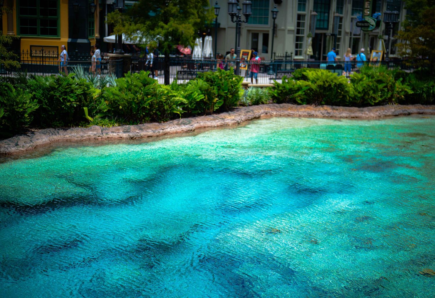 Crystal clear turquoise water at a tropical resort lagoon with lush green foliage and visitors along the shoreline.