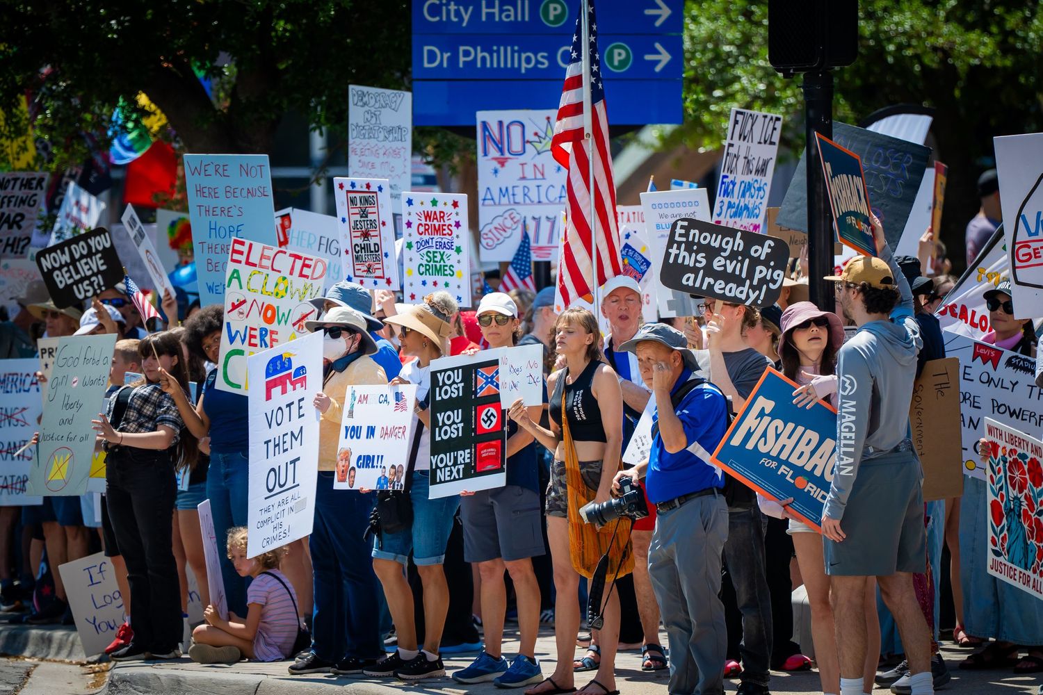 Protesters gather holding signs and an American flag at a political demonstration on a sunny day.